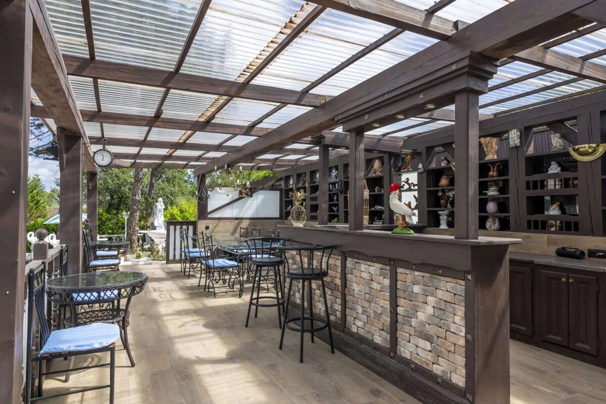 A rustic outdoor patio with a clear corrugated roof, wooden beams, tables, chairs, and a bar featuring decor, a rooster statue, and distant greenery.