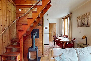 Cozy rustic interior featuring a wooden staircase with rope railing, a wood stove, a dining area with red chairs, and a white sofa.
