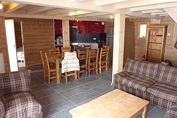 A rustic, open-plan chalet interior featuring wood-paneled walls, exposed beams, a red kitchen, wooden dining set, and plaid sofas on a dark tiled floor.