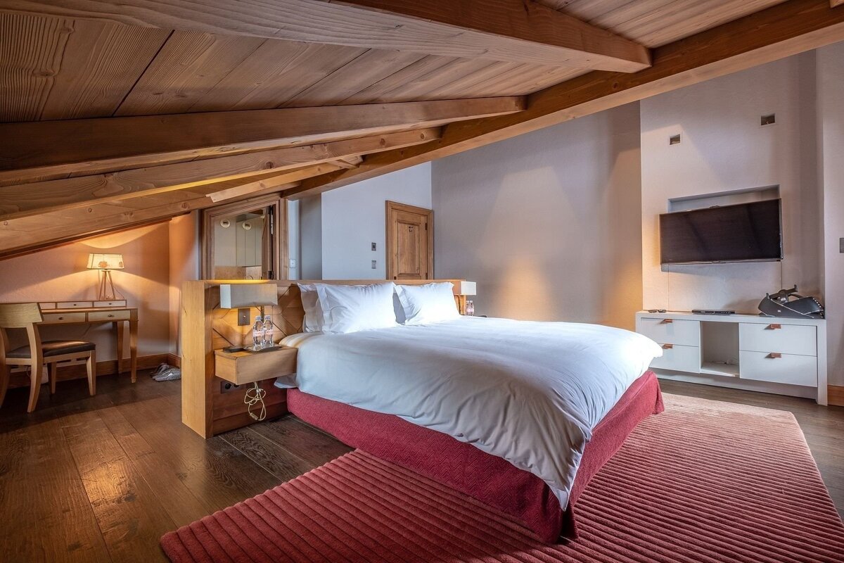 A cozy attic bedroom featuring a large bed with a red base, white duvet, and a matching red rug on dark wood floors. A wooden beamed ceiling adds character.