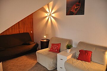 A cozy living room features a dark futon, two light armchairs with red pillows, wood paneling, and warm lighting from a wall sconce and lamp.