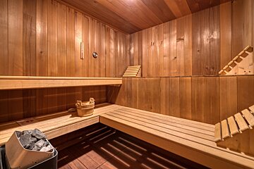 A cozy, warm wooden sauna with multi-level benches, a water bucket, and a heater with hot stones. The walls and ceiling are wood-paneled.