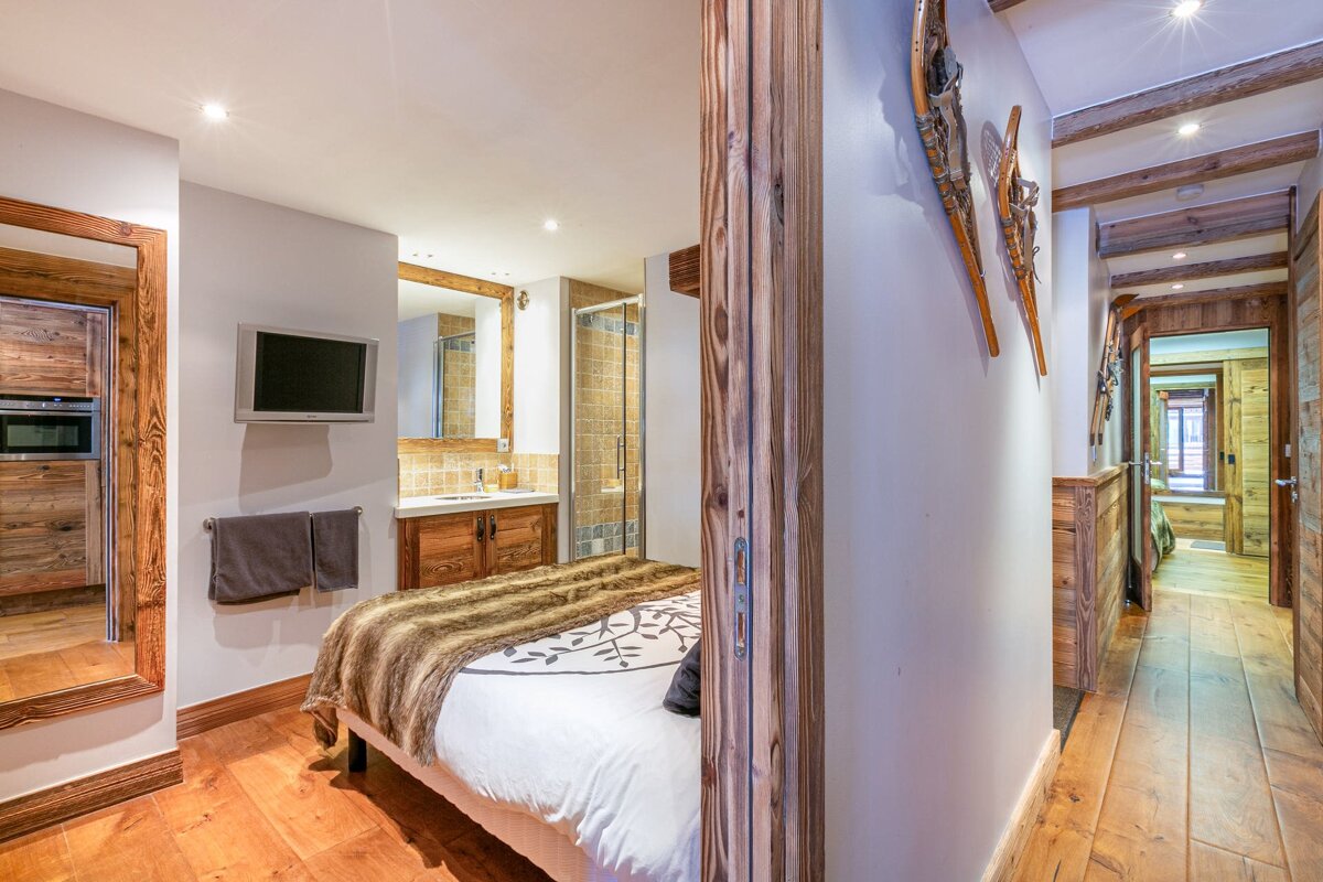 Cozy alpine bedroom with a bed, TV, integrated bathroom, and a rustic wooden hallway adorned with snowshoes. Warm wood tones dominate.