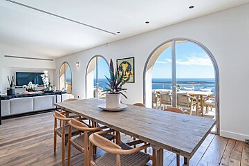 Bright dining room with a long wooden table and chairs, connected to a living area. Arched windows offer stunning ocean views from a balcony.