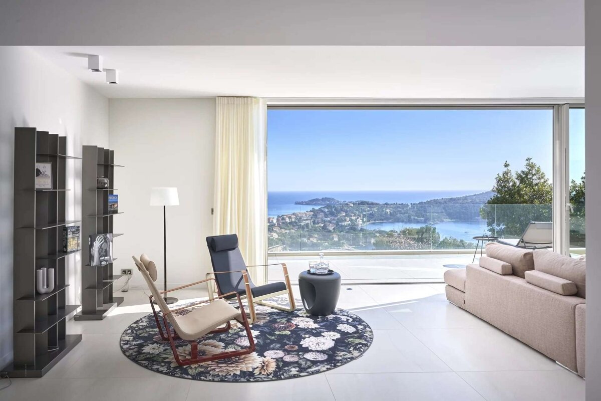 A modern living room with stunning sea views through floor-to-ceiling windows, featuring stylish chairs, a floral rug, sofa, and bookshelves.