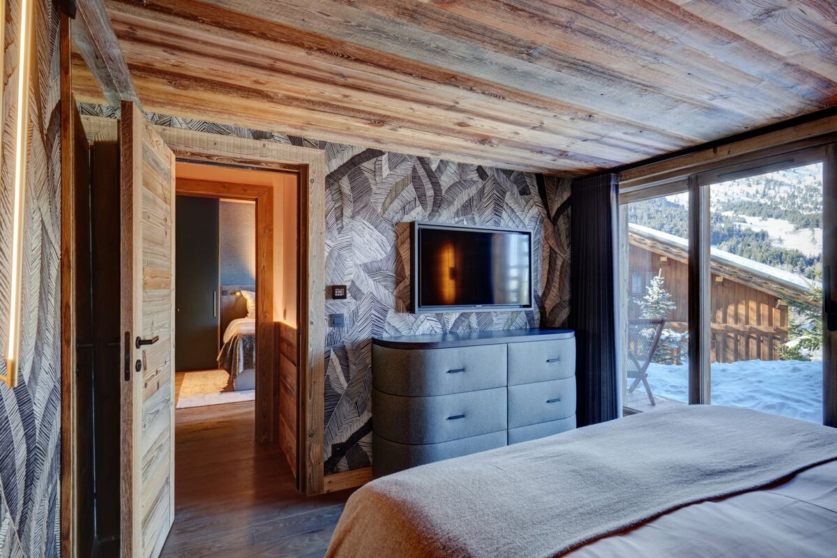 A rustic-chic bedroom with a wood ceiling, leaf wallpaper, TV, dresser, and a large window showcasing a snowy mountain view. An open doorway reveals another room.