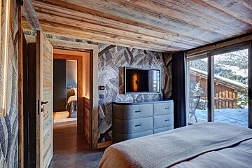 A rustic-chic bedroom with a wood ceiling, leaf wallpaper, TV, dresser, and a large window showcasing a snowy mountain view. An open doorway reveals another room.