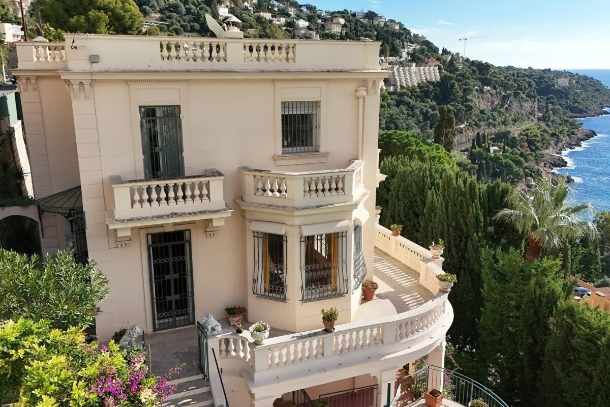 An elegant cream-colored coastal villa with balustraded terraces on a lush green hillside overlooking the blue sea.