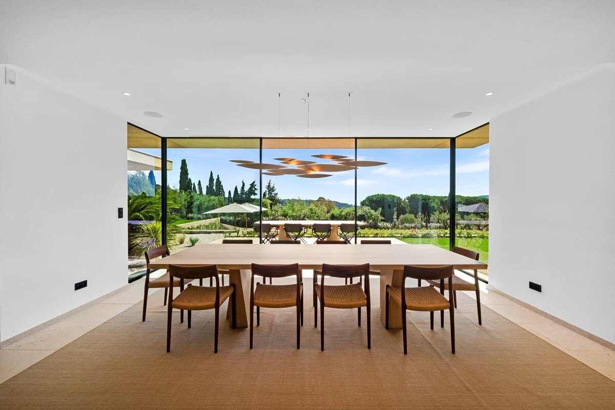 A modern dining room with a long wooden table, chairs, and floor-to-ceiling windows showing a scenic backyard.