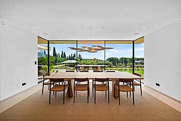 A modern dining room with a long wooden table, chairs, and floor-to-ceiling windows showing a scenic backyard.