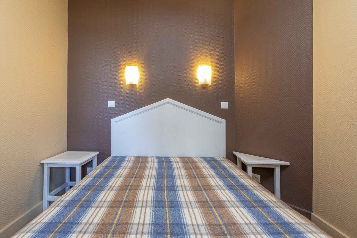 A small bedroom with a single bed, plaid blanket, white headboard, two nightstands, and two lit wall sconces against textured brown and beige walls.
