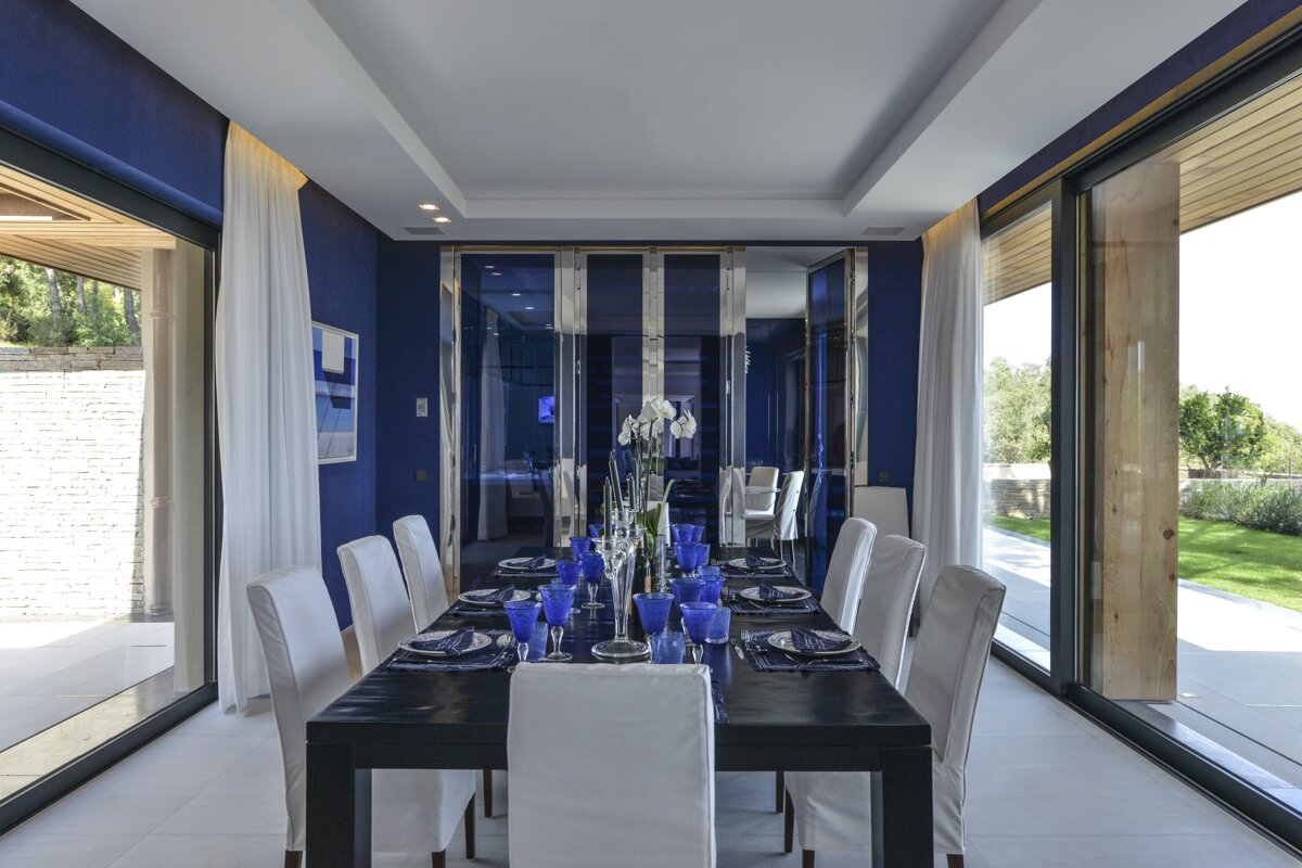 A modern, elegant dining room with deep blue walls, a long black table set with white chairs and blue tableware, flanked by large glass doors to the outdoors.