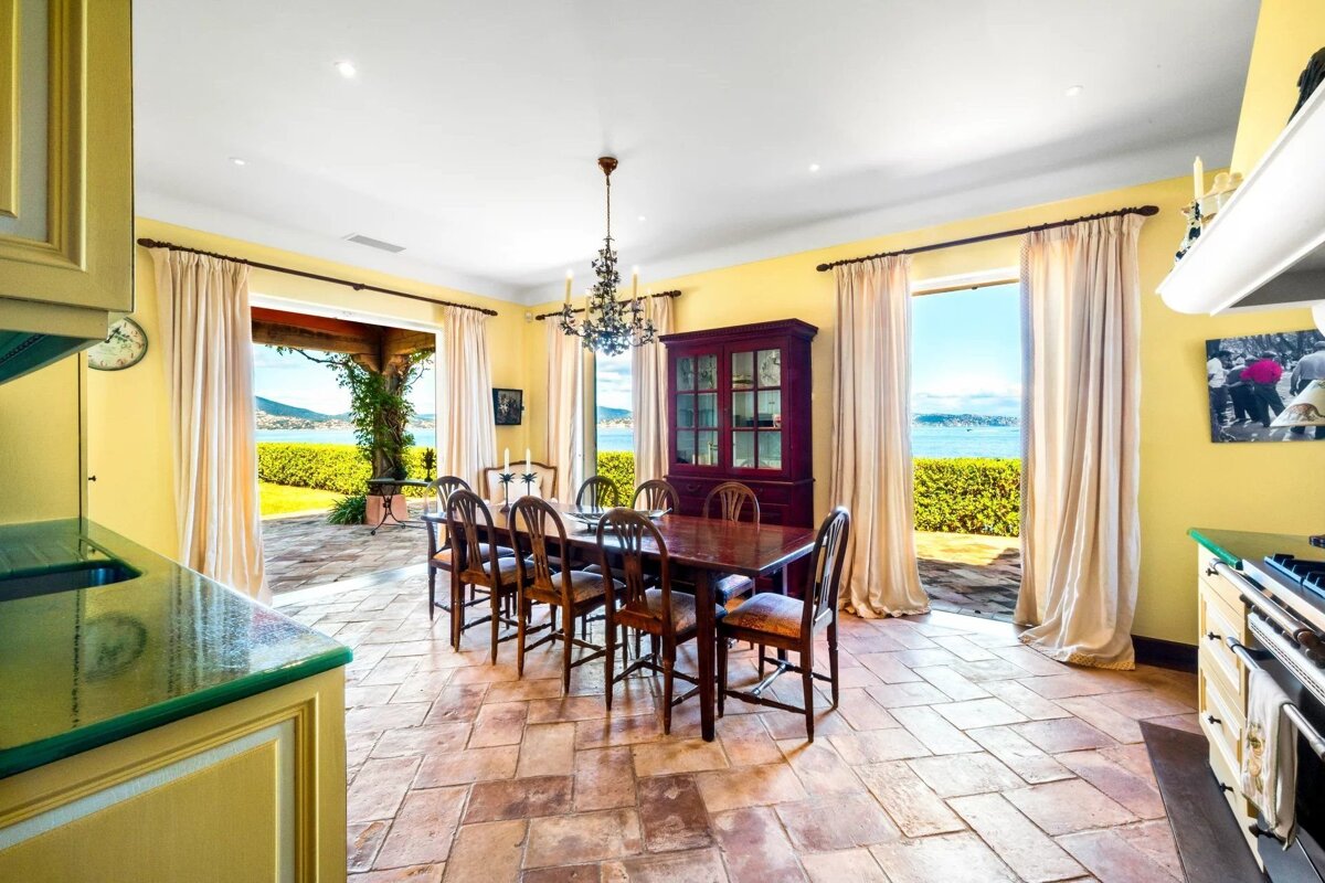 A bright dining room with a long wooden table, tiled floor, and open doors leading to a patio overlooking the sea.