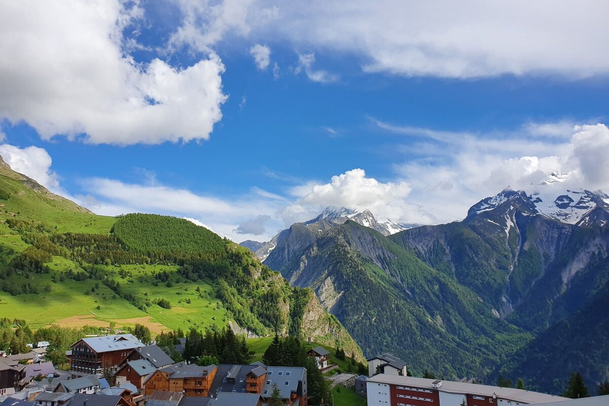 A picturesque mountain village nestles among lush green slopes and towering, snow-capped peaks under a bright blue, cloudy sky.