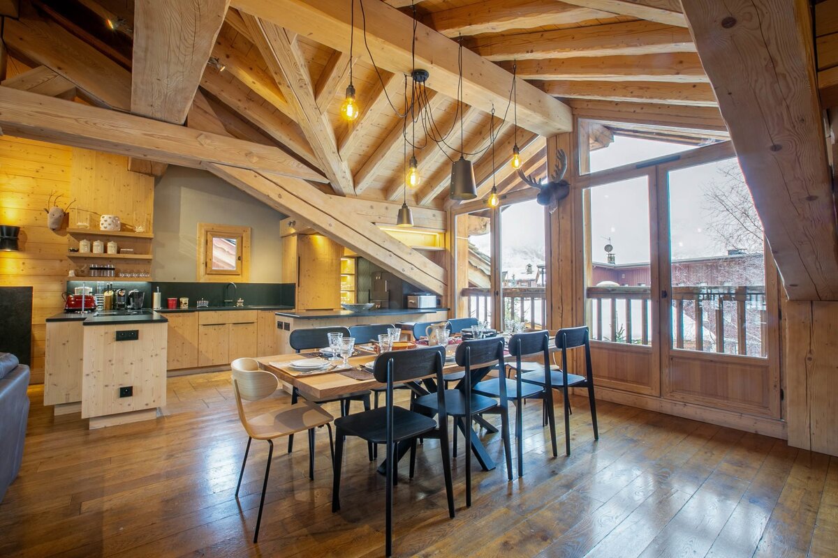Cozy wooden chalet interior with kitchen, dining area, exposed beams, and large windows revealing a snowy mountain view.