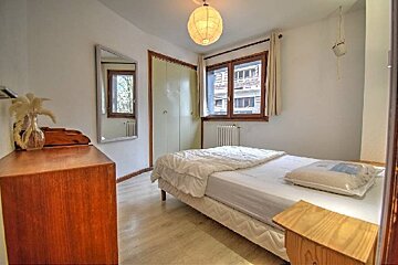 A brightly lit bedroom featuring a bed, wooden furniture, a full-length mirror, a window, and a spherical pendant lamp.