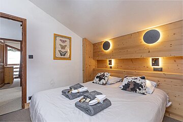 A modern bedroom features a wooden headboard with circular and reading lights, a white bed with cow-print pillows, and towels. A butterfly print hangs on the white wall.