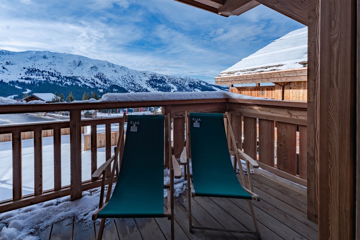 Two green lounge chairs on a snowy wooden balcony with a stunning view of snow-capped mountains.