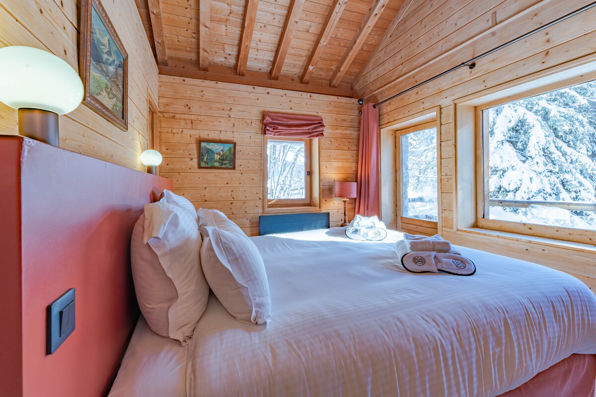 Cozy wooden cabin bedroom featuring a neat bed and large windows with a view of a snowy pine forest.