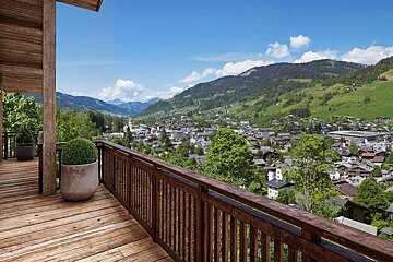 A wooden balcony offers a scenic view of a mountain village nestled among green hills under a clear blue sky.