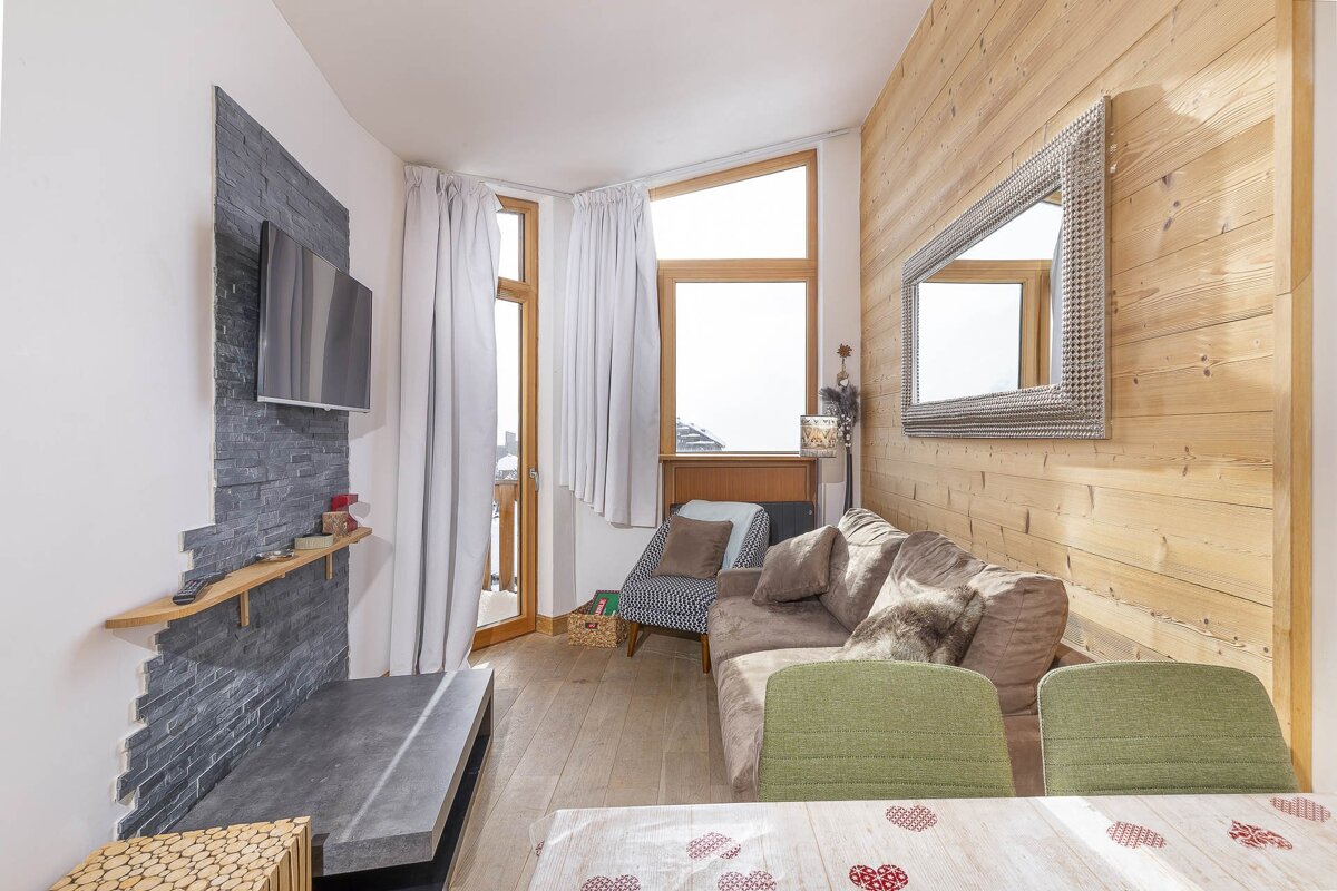 A cozy, modern chalet living room with wood paneled walls, a gray stone accent wall with TV, comfy sofa, and dining area.