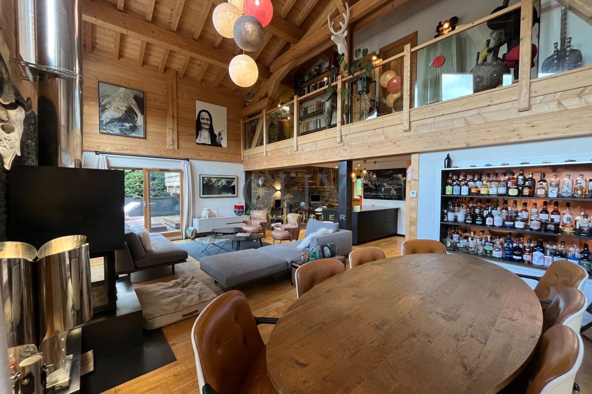 A spacious wooden chalet interior with a large dining table, cozy living room, mezzanine loft, and a well-stocked bar shelf.