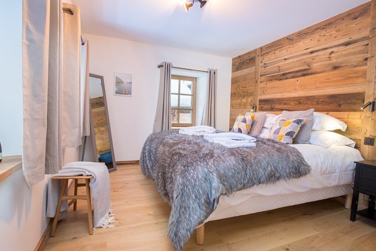 A cozy bedroom with a rustic wooden accent wall, light wood floors, a standing mirror, and a bed topped with a plush grey faux-fur throw.