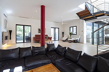 A bright, modern living room featuring a black sofa, red pillar, white recliners, large windows, and a contemporary staircase.