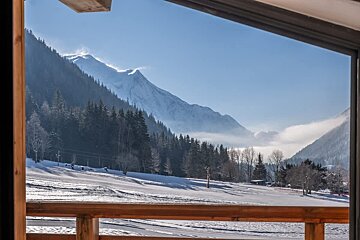 A scenic winter view of snow-covered mountains and pine forests, framed by a wooden balcony under a clear blue sky.