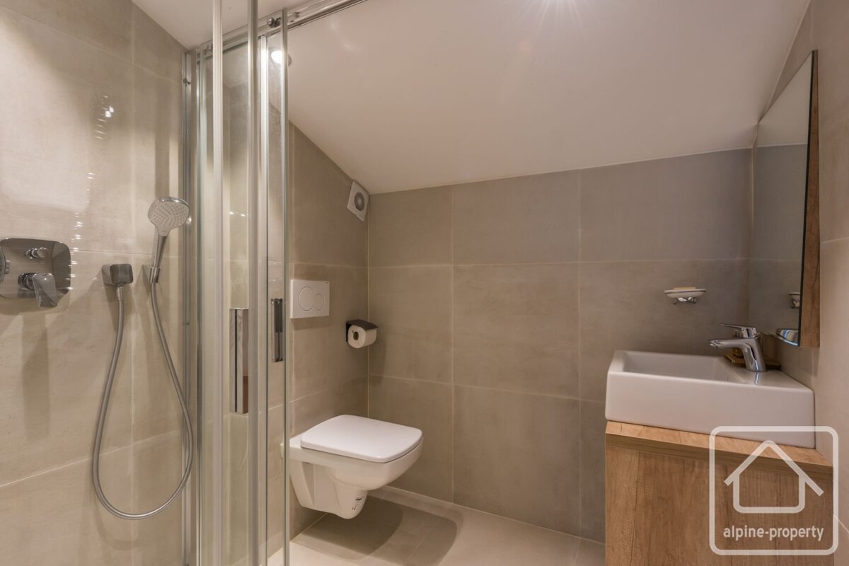 A modern bathroom with a glass shower, wall-mounted toilet, and white sink on a wooden vanity, featuring neutral tiled walls.