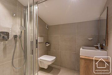 A modern bathroom with a glass shower, wall-mounted toilet, and white sink on a wooden vanity, featuring neutral tiled walls.