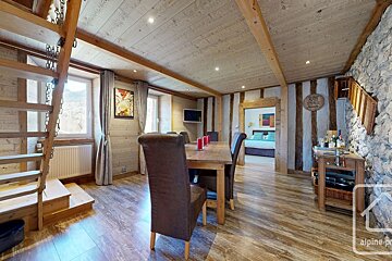 Rustic dining room with wood paneling, exposed beams, and a stone wall. Features a dining table, chairs, ladder stairs, and a bedroom view.