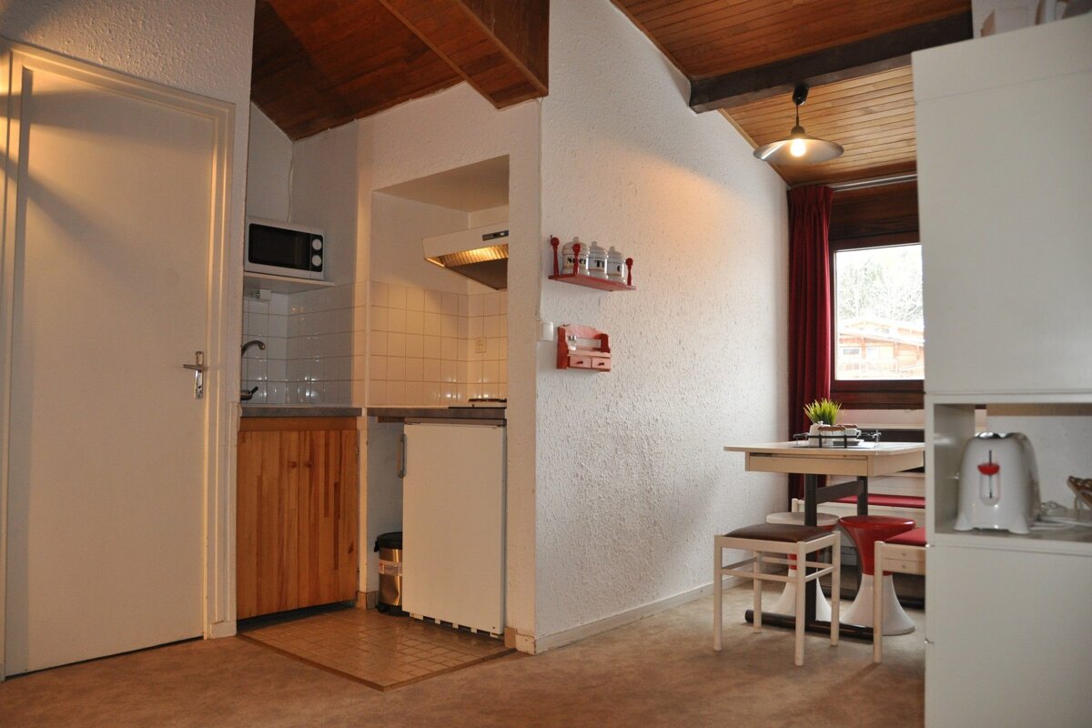 A compact kitchen and dining nook with white textured walls, rustic wooden ceiling beams, and modern appliances. Red accents add warmth to the space.