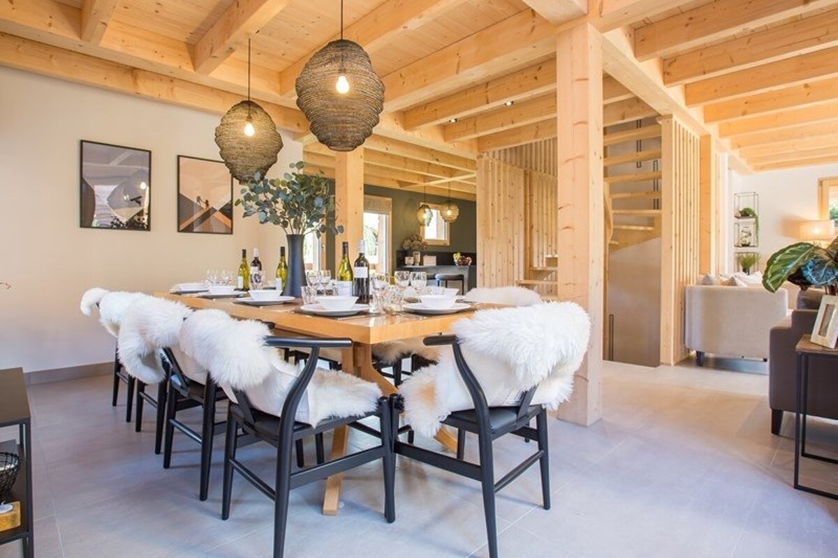 A modern dining area featuring a wooden table, chairs with white sheepskin throws, woven pendant lights, and a light wood beamed ceiling.