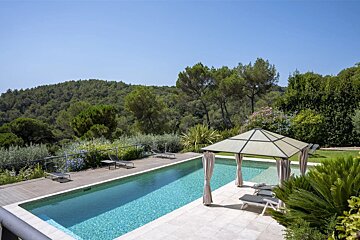 A luxurious swimming pool with a gazebo and loungers, overlooking lush green forested hills under a clear blue sky.