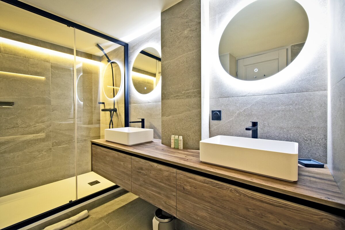 Modern bathroom with a wooden double vanity, white vessel sinks, backlit circular mirrors, and a glass-enclosed shower.
