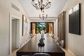 A long dining room features a dark wooden table, clear chairs, two chandeliers, and modern art. Glass doors reveal a lush garden.