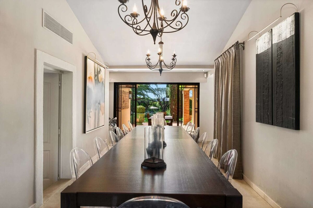 A long, elegant dining room with a dark wooden table, clear chairs, ornate chandeliers, and modern art. Glass doors open to a lush outdoor patio.