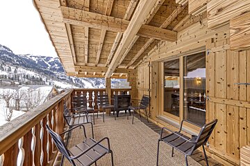Cozy wooden chalet balcony with chairs and a fire pit, offering stunning snowy mountain views and a rustic ambiance.