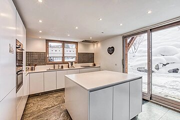 Sleek white kitchen with a central island, tiled floor, and a large glass door leading to a snow-covered outdoor scene.