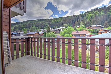 A view from a wooden balcony overlooking a charming alpine village nestled against a lush green forest under a cloudy sky.