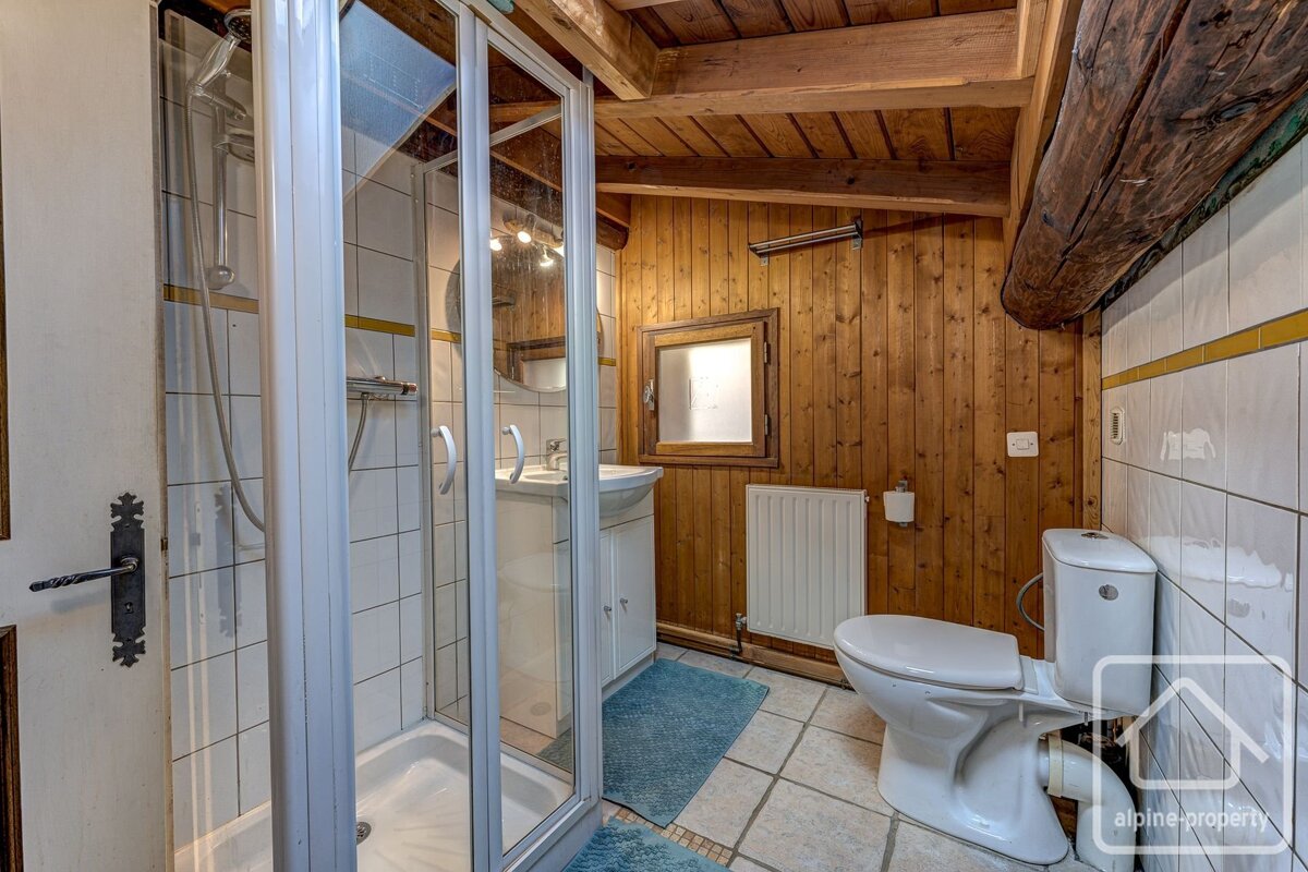 A rustic bathroom with wood-paneled walls, white tiled sections, a glass shower, toilet, sink, and exposed wooden ceiling beams.