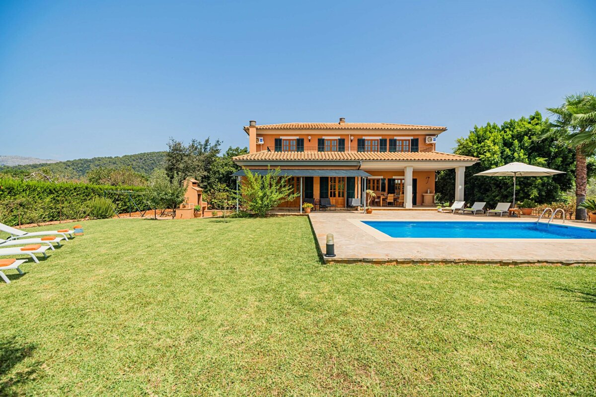 An orange villa with a swimming pool, spacious green lawn, and lush hills under a clear blue sky.