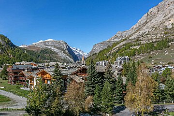 An alpine village with traditional chalets and a church spire nestled in a valley, framed by snow-capped mountains under a clear sky.