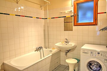 A brightly lit bathroom with white tiles, a bathtub, sink, washing machine, and a wooden-framed window. A colorful stripe decorates the wall.