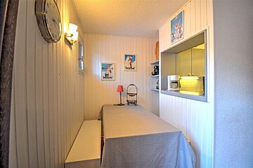 A cozy, narrow dining area featuring white paneling, a long table with bench seating, ski posters, and a kitchen pass-through.