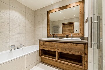 A rustic-modern bathroom featuring a wooden double vanity with two sinks, a large wood-framed mirror, a bathtub, and tiled walls.
