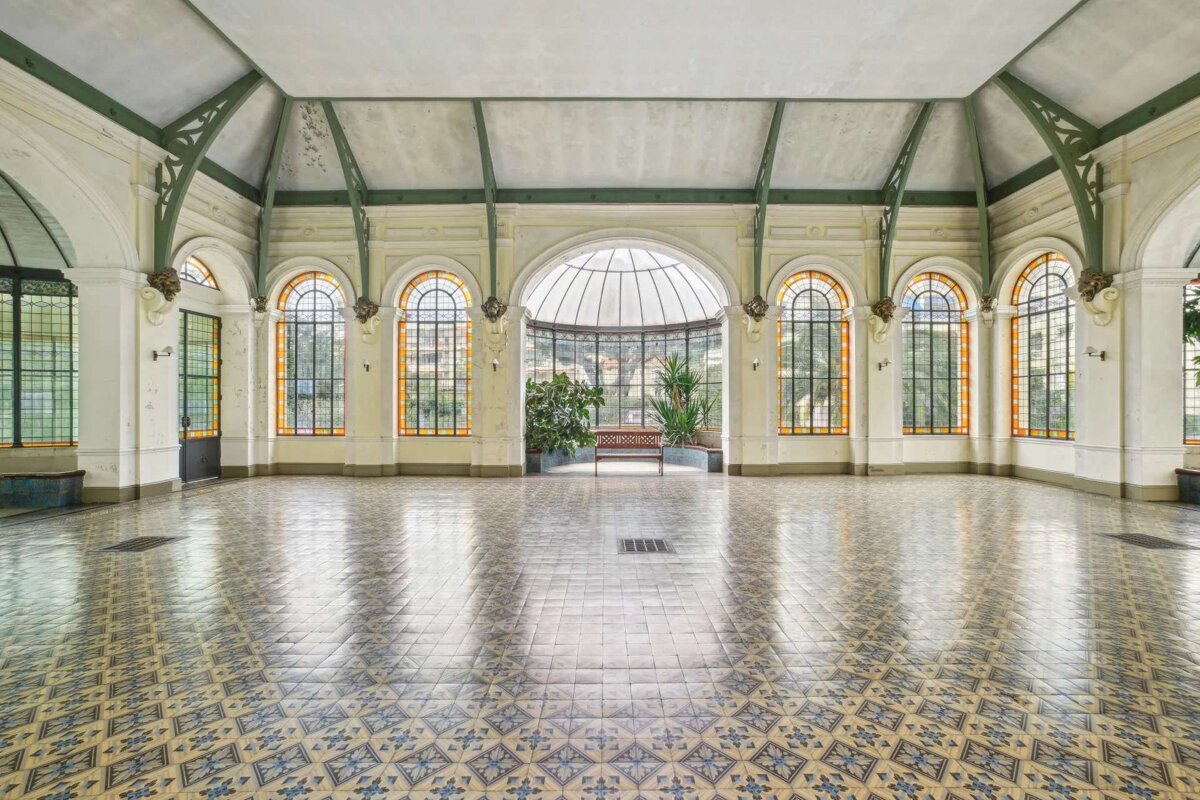 A grand, empty hall with patterned tiles, arched windows with stained glass accents, and a glass-domed end featuring plants and a bench.