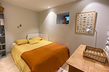A cozy bedroom features a bed with yellow and orange linens, a grey bookshelf, an insect chart, and a wooden desk with a lamp.