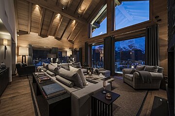 A warm, luxurious chalet living room with vaulted wooden ceilings, cozy seating, and large windows revealing a beautiful, snowy mountain evening view.